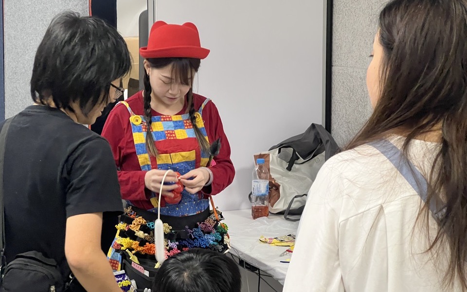 一位戴紅帽、穿彩色吊帶褲的藝人在活動現場的工作枱前即場製作，旁邊有家長與小朋友等候